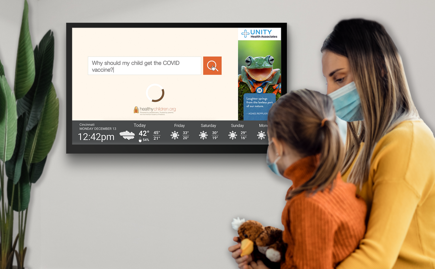 A young child and mother sit in a doctor's office wearing masks looking at a screen on the wall with information about the COVID-19 vaccine for children