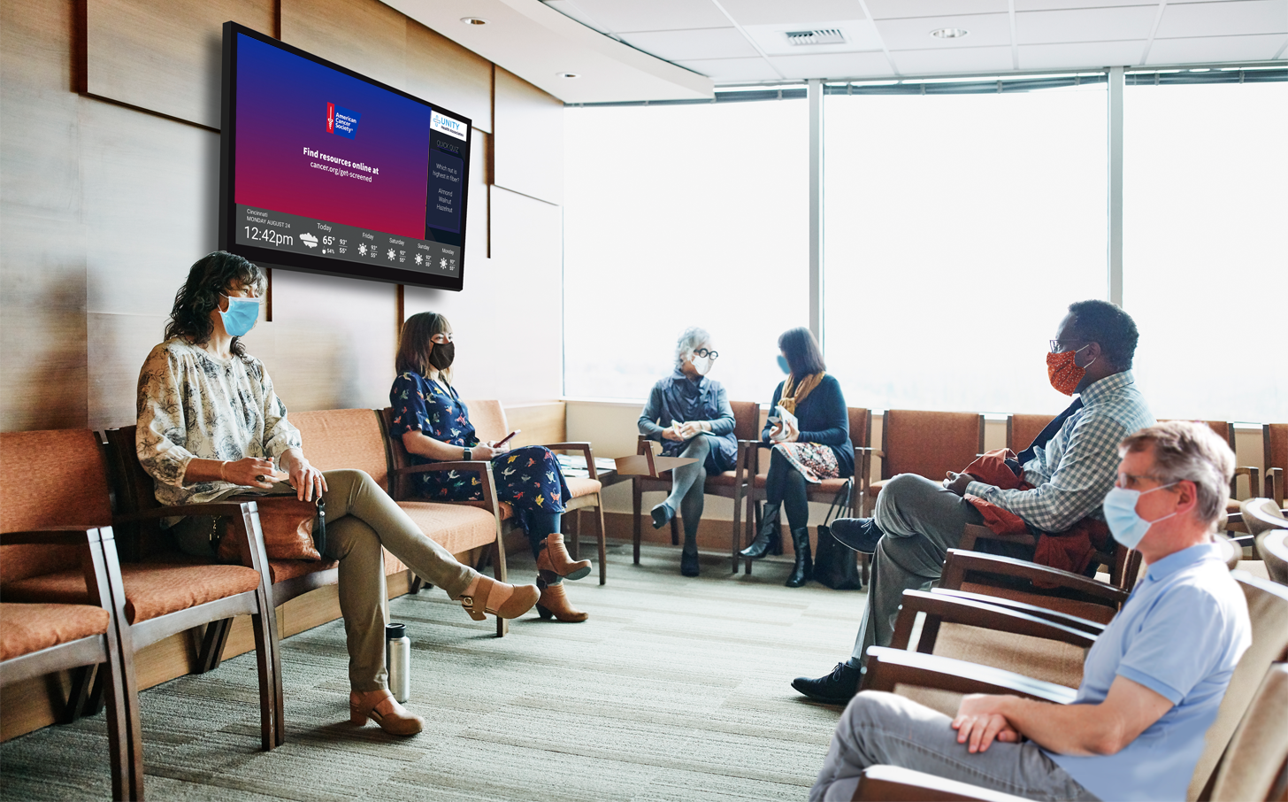 Patients sit in a waiting room observing American Cancer Society content about the importance of regular screenings.
