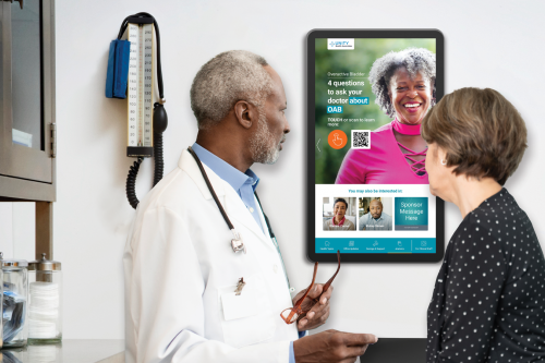 A mature male physician reviews content about overactive bladder on a wall-mounted touchscreen in an exam room with a middle-aged female patient. 